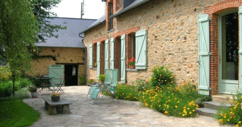 grande longère en pierre, contours de portes et fenêtres en brique rouge, volets et huisseries en bois vert clair, terrasse fleurie en ardoise