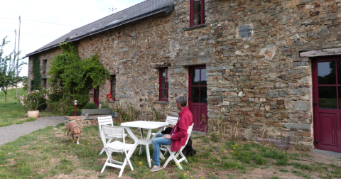 longère en pierre avec huisseries bordeaux, petite terrasse en herbe avec homme et ses chiens assis à une table de jardin blanche