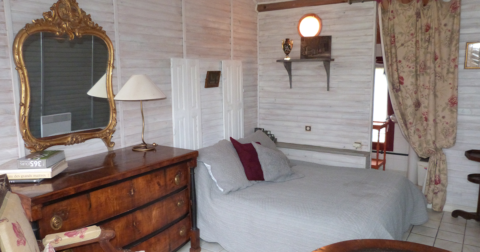 petite chambre avec murs en bois blancs, carrelage, lit double avec couverture grise et commode et miroir de style ancien