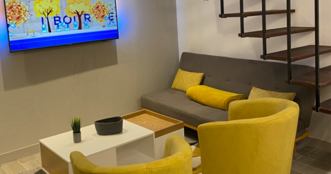 coin salon avec canapé banquette grise fauteuils jaune, table basse en bois et résine blanche, écran plat suspendu au mur