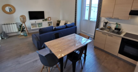 appartement avec cuisine aménagée en bois, petite bale en bois et chaises noires, coin salon télé avec canapé 3 places bleu