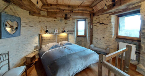 Chambre ronde en pierre avec poutre en bois, lit double aux draps gris et tête de lit en bois de récupération