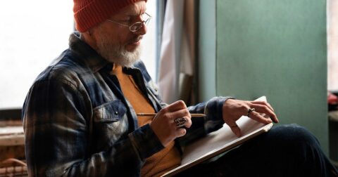 homme d'âge mûr avec lunettes, chemise à carreaux et bonnet rouge en train de réfléchir avant d'écrire