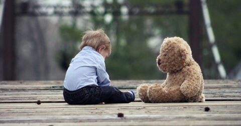 très jeune garçon assis sur dehors sur des planches en bois faisant face à son ours en peluche