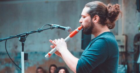 Un homme barbu et cheveux longs relevés de profil jouant de la flûte devant un micro