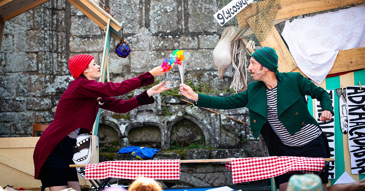 deux comédiens, l'une en rouge et l'autre en vert, chacun d'un côté d'une table, se tendent des objets insolites