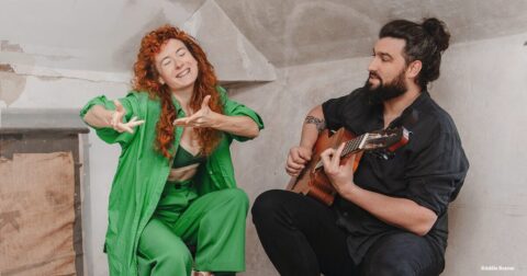 une femme en vert chantant et un homme en noir jouant de la guitare dans un grenier