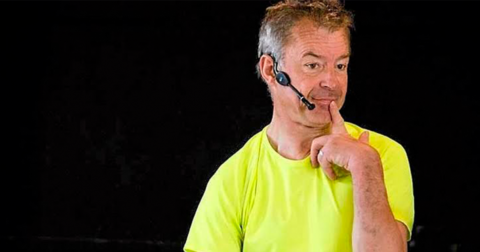 un homme sur scène avec t-shirt jaune fluo, une oreillette et un doigt posé sur la bouche